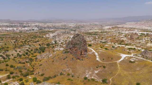 Goreme, Nevsehir, Turkey. A solitary volcanic tuff monolith rises above the arid Goreme Valley, an aerial panoramic view over eroded cliffs and scattered greenery under clear summer skies. Aerial Vie