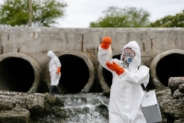 Scientists in hazmat suits collect water samples near concrete drainage pipes discharging polluted wastewater, testing contamination and environmental hazards at an outfall site. © akarawit