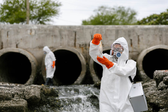 Scientists in hazmat suits collect water samples near concrete drainage pipes discharging polluted wastewater, testing contamination and environmental hazards at an outfall site.