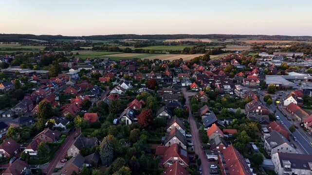 Red-roofed houses spread across quiet German suburb at sunset, narrow streets wind between dense tree canopy as open farmland stretches into calm evening distance. Aerial drone wide shot. Quiet scene