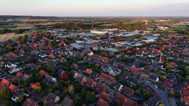 Dense red-roofed town spreads across gentle hills in Germany, industrial buildings and church spire rise among tree-lined streets under late summer evening light. Aerial drone flyover shot. Sunrise.