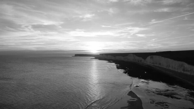 Aerial drone view showing chalk cliffs, coastline and calm sea with sunlight reflecting across water beneath cloudy sky in Brighton, England, United Kingdom