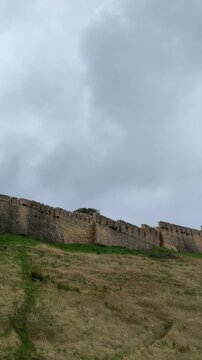 Crenellated stone fortress wall rises from steep grassy slope beside rounded tower, with weathered masonry, sparse vegetation, and soft grey cloud cover creating subdued historic atmosphere