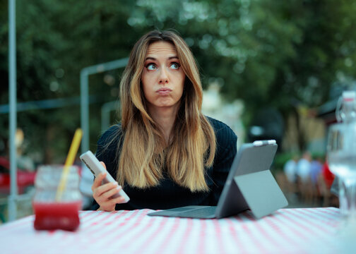 Stressed Young Woman With Puffed Cheeks Working On Her Tablet. An overwhelmed girl holds her phone while multitasking with a tablet indoors