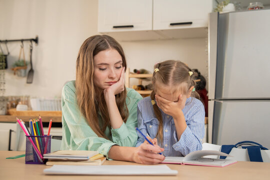 Mother gently watching her stressed daughter covering face while doing difficult homework. Mother sitting at kitchen table supporting daughter who feels frustrated and overwhelmed by home schooling.