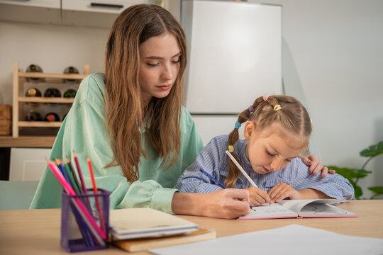 Little girl excitedly talking while writing homework with mother's supportive hand on her shoulder. Young girl enjoying homeschooling study time with her mother providing guidance and care at home.