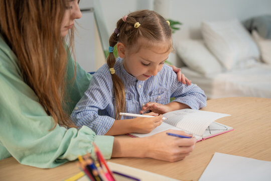 Little girl excitedly talking while writing homework with mother's supportive hand on her shoulder. Young girl enjoying homeschooling study time with her mother providing guidance and care at home.