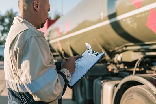 Person in high-visibility coveralls performing a safety audit with a clipboard beside a polished chemical road tanker, with copy space