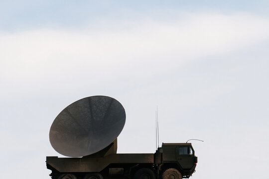 Large parabolic dish antenna on a heavy olive green truck, featuring a weathered mesh reflector and muddy tires for defense technology and communications infrastructure, with copy space