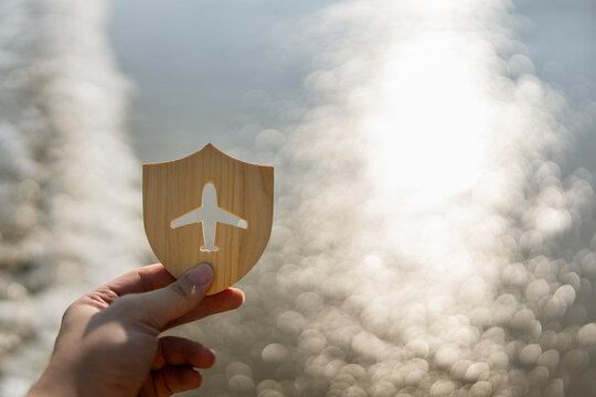 Hand holding wooden shield with airplane icon against blurred sea bokeh background representing travel insurance protection flight safety tourist security concept.