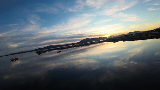 Small boats rest near Muelle de Pescadores Artesanales, with mirror still water reflecting a vivid sunset sky near Puerto Natales, Chile, FPV aerial