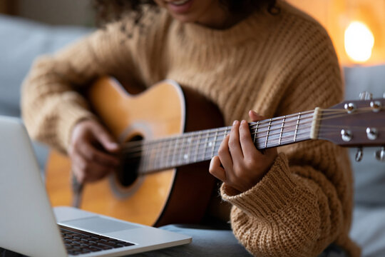 Cozy room with warm lighting. Person plays guitar with focus and joy. Laptop nearby suggests learning or streaming. Comfortable sweater adds homely atmosphere