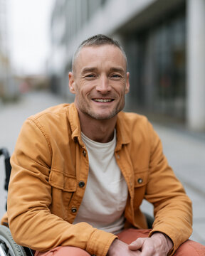 Man in orange jacket smiles warmly in city setting. He sits comfortably in wheelchair with relaxed posture. Background features blurred urban architecture and sidewalk