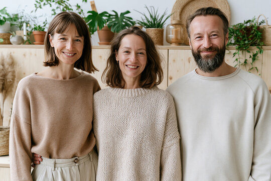 Three smiling people pose warmly beside plants. Wooden shelves hold potted greenery and dried flowers. Natural light enhances cozy, earthy tones of their outfits. Their expressions radiate joy