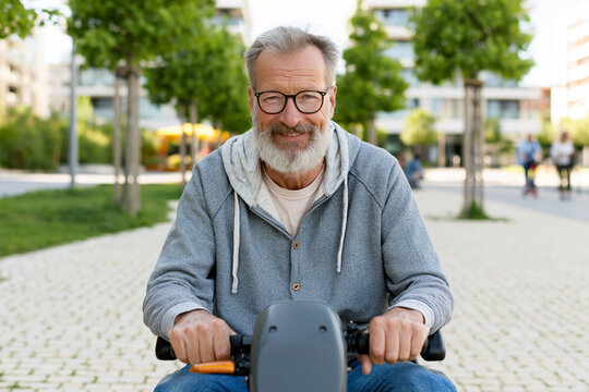 Grey-haired man smiles while riding electric scooter. Wears glasses, hoodie, and jeans. Urban park setting with trees and buildings. Background shows blurred pedestrians and greenery
