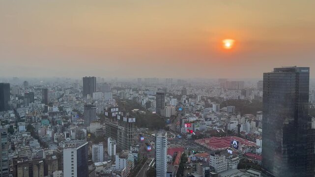A high view of the city skyline during sunset. Many tall buildings and houses fill the large urban area under a warm orange and hazy sky, seen from a high place in the late afternoon.