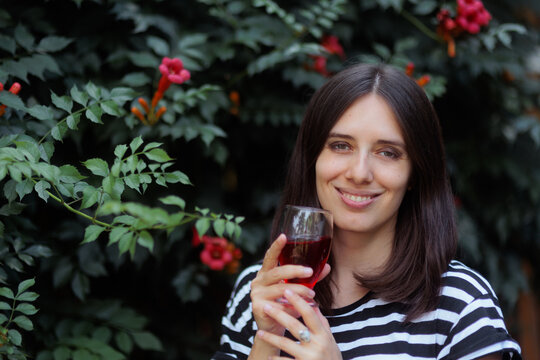 Beautiful Young Woman Smiling And Holding A Glass Of Red Wine. A happy attractive lady enjoys drinking her beverage outdoors near some flowers