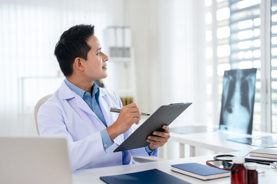 Young doctor  filling paper medical records. Practitioner in white coat doing paperwork at workplace with laptop, writing notes, preparing documents, reports, prescription.