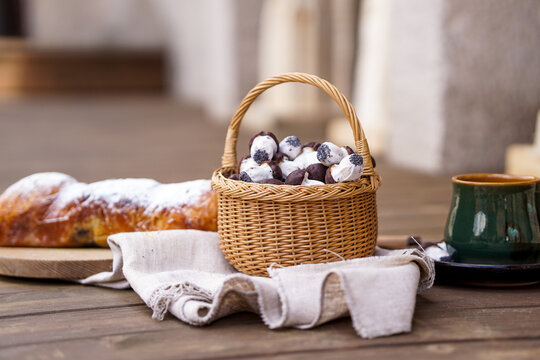 A rustic table setup featuring a powdered sweet bread roll on a wooden board, a green ceramic cup, and a basket full of mushroom-shaped chocolate cookies.