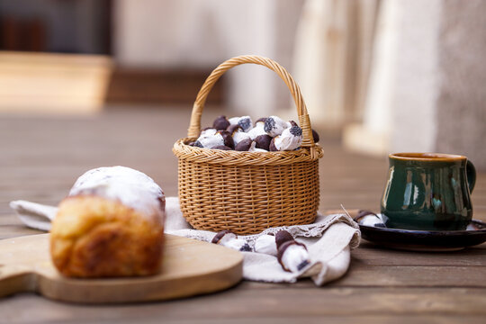 A rustic table setup featuring a powdered sweet bread roll on a wooden board, a green ceramic cup, and a basket full of mushroom-shaped chocolate cookies.