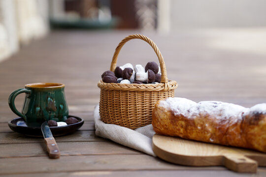 A rustic table setup featuring a powdered sweet bread roll on a wooden board, a green ceramic cup, and a basket full of mushroom-shaped chocolate cookies.