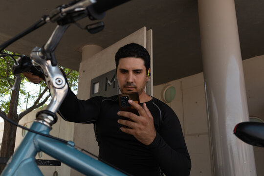 Male cyclist stopping during urban commute to check smartphone, using wireless earbud for navigation or communication while standing near his bicycle