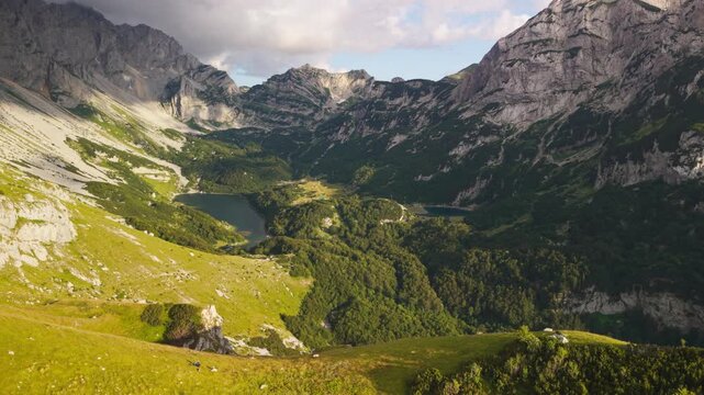 Drone reveal shot pulling back from hikers resting on grass to a wide aerial view of Skrcka Valley and Bobotov Kuk peak in Durmitor National Park, Montenegro