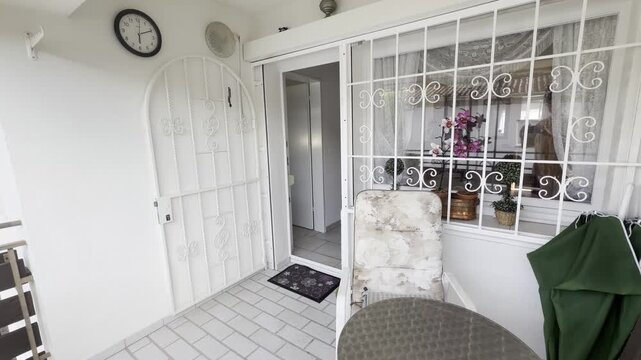 A slow pan across a bright residential balcony featuring an open white wrought-iron gate, a round metal table, a garden chair, a folded patio umbrella, and a window overlooking the home interior.