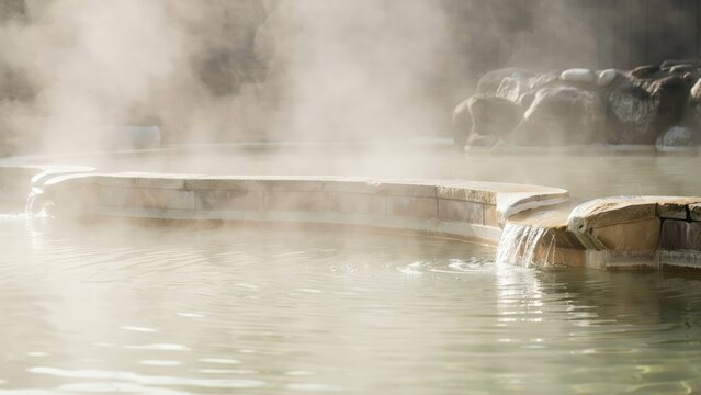 Steam rises from a natural hot spring pool with stone architecture and flowing water during daylight. Relaxing wellness concept for spa advertisements and travel promotion.