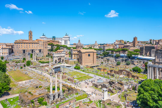 Tourists walk among ancient ruins at the Roman Forum in Rome Italy. The historic site shows remains of buildings and columns under a blue sky with few clouds.