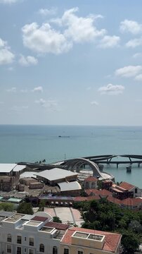 4k footage of panoramic view of a coastal town by the blue sea. Colorful buildings and a large arena sit near the water, with a cable car and boats moving along the clear ocean under a sunny sky