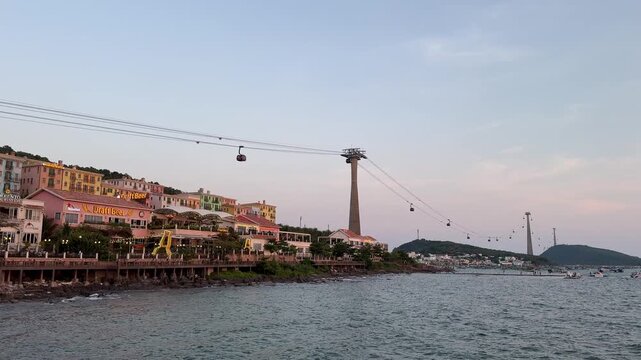 4k footage of cable cars moving over a calm sea near a coastal town. Colorful buildings and restaurants line the shore with glowing lights as evening falls over the peaceful water.
