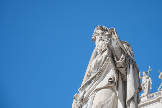 Saint Paul stands tall at St Peters Basilica in Vatican City. The statue shows him with a book in one hand, under a clear blue sky. Visitors admire the fine details of the sculpture.