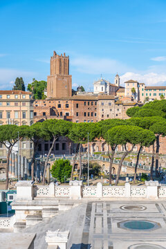 Visitors explore the ancient Trajan Forum in Rome. The site shows historic buildings and greenery under a blue sky. The lively scene captures the essence of Roman history and culture.