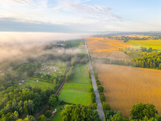 Obraz premium Aerial view shows fields and road stretching across the landscape. Fog covers parts of the land creating a soft barrier. Trees line the edges, and sunlight illuminates the scene.