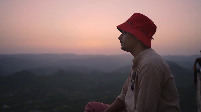 Person in red bucket hat watching sunset over mountains in India