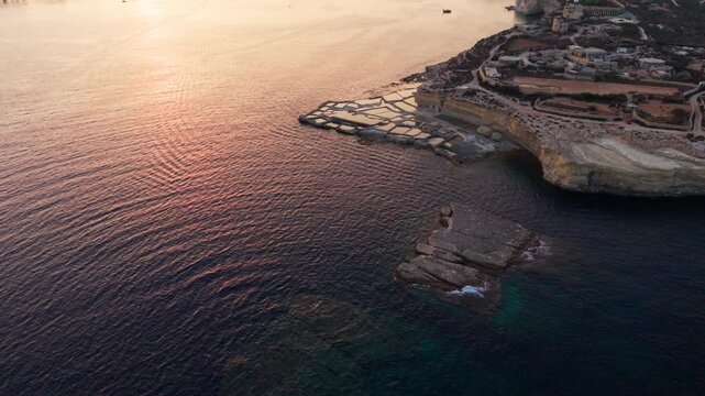 Delimara salt pans filled with water reflecting the sunset, captured from a 45-degree aerial drone perspective with a slow forward flight over the calm Mediterranean Sea.