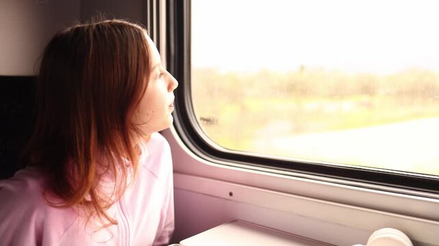 A girl sits by a train window and gazes outside. She watches fields and trees pass by as the train moves. It is a bright day and she seems thoughtful while enjoying the journey.