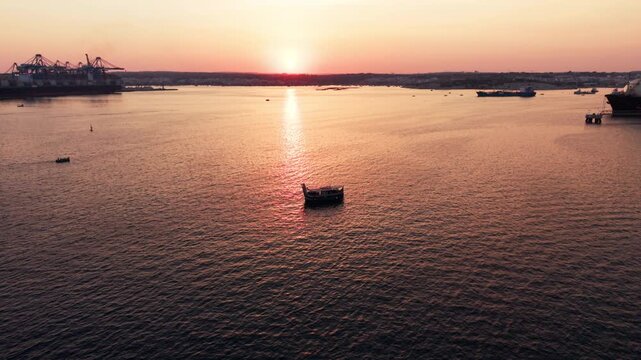 Sunset over the Mediterranean Sea with a silhouetted Maltese luzzu and Malta Freeport visible in the distance, captured in a wide-angle drone shot with slow orbit over calm water.