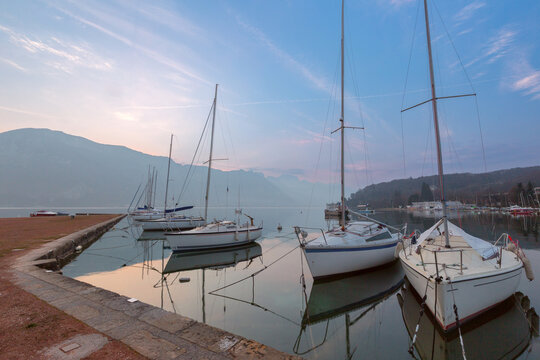 Sailboats reflected in the calm water of Lake Annecy at sunrise with mountains in the background, France.