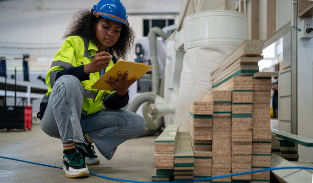 Warehouse Inspection: A diligent black female worker in a hard hat and safety vest crouches by a stack of lumber, meticulously recording details on a clipboard. Multiracial woman worker.
