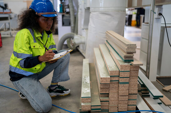Warehouse Inspection: A diligent black female worker in a hard hat and safety vest crouches by a stack of lumber, meticulously recording details on a clipboard. Multiracial woman worker.