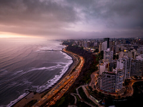 Aerial view of the coastal skyline of Lima at dusk, featuring high-rise buildings, a busy highway, and Pacific Ocean waves crashing against the shore in Lima, Callao Region, Peru.