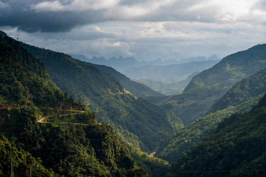 Aerial view of the winding Ha Giang Loop road cutting through lush green mountains and deep valleys under a dramatic cloudy sky in Dong Van, Tuyen Quang, Vietnam.