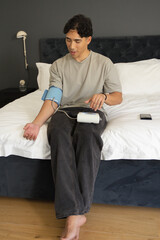 Fototapeta premium Asian man sitting on bed in bedroom, checking blue blood pressure cuff and monitor with smartphone