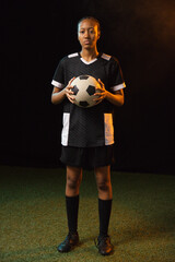 Fototapeta premium African adult woman standing on green turf studio-lit holding black-white soccer ball in black kit
