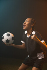 Fototapeta premium African American woman soccer player shouting, holding ball in black-white kit in studio on turf