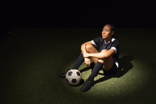 African American female soccer player sitting on turf in spotlight, wearing black kit, ball nearby