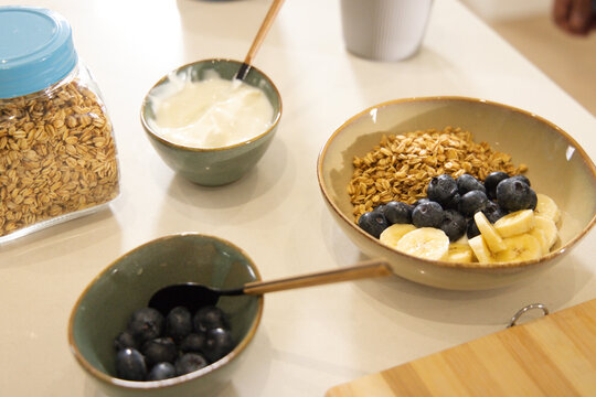 Granola bowl sitting on light counter with blueberries, banana, yogurt, oats jar and brass spoons