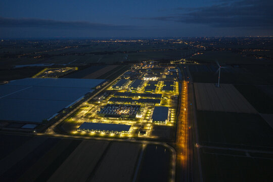 Aerial view of an illuminated hyperscale AI data center at night, featuring rows of server buildings, cooling infrastructure, and surrounding dark fields.